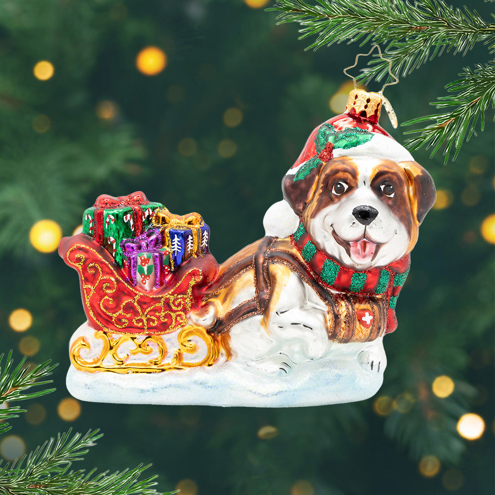 Glass Saint Bernard ornament wearing a Santa hat and pulling a red sleigh filled with gifts.