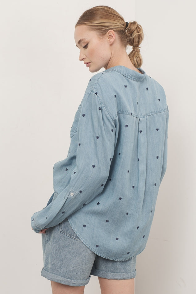 Woman wearing an oversized light blue chambray shirt with dark heart embroidery and denim shorts.