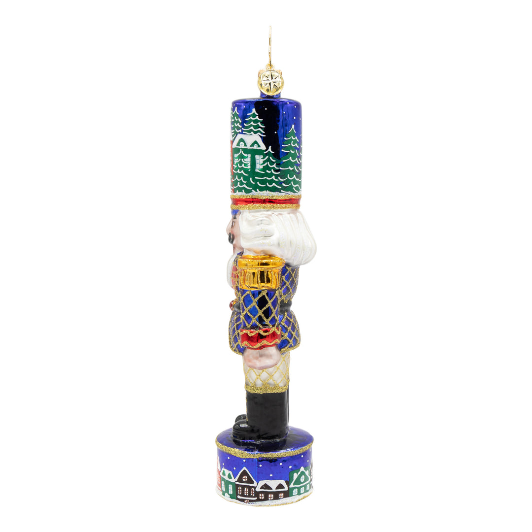 Royal blue nutcracker ornament with a snowy village scene on his hat and base.