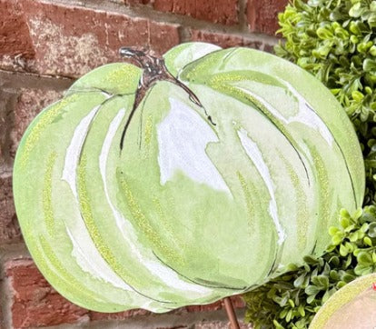 Light green painted pumpkin decoration with white highlights and a brown stem against a brick wall.