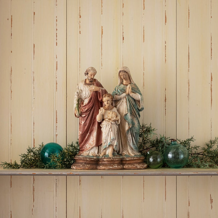 A 16-inch resin Holy Family statue with an aged, antique finish on a shelf with green ornaments.