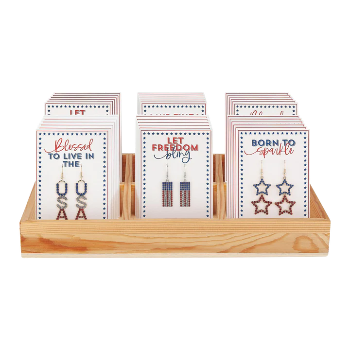 Three displays of patriotic USA-themed earrings on cards in a wooden tray. Earrings include USA letters, flag rectangles, and stars.