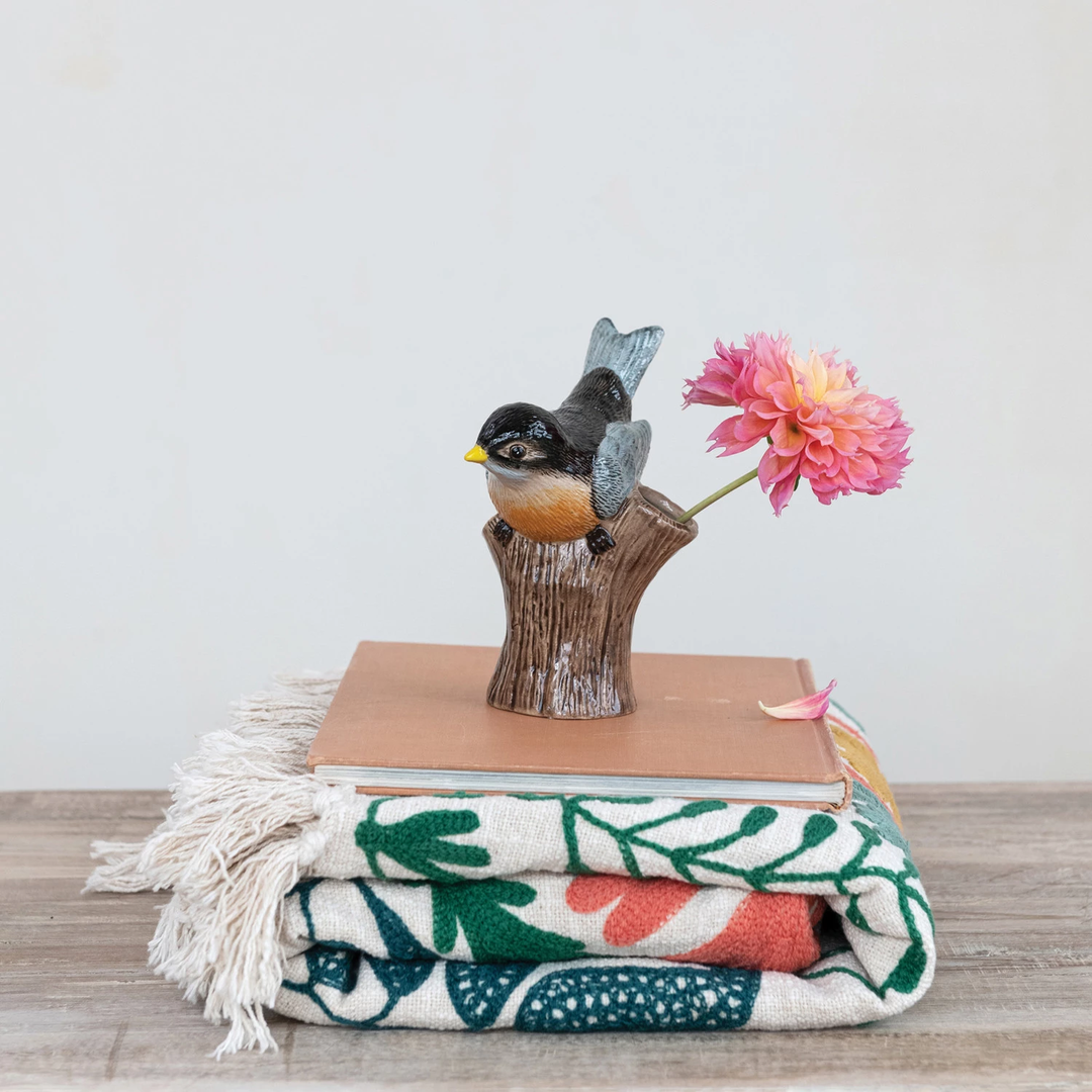 Hand-painted stoneware bird on a log-shaped vase holding a pink flower, atop a book and blanket.