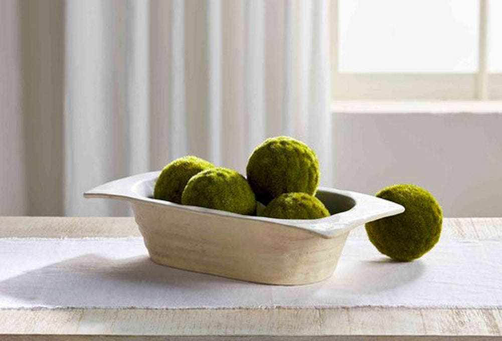 White stoneware dough bowl on a table runner filled with decorative green moss balls.