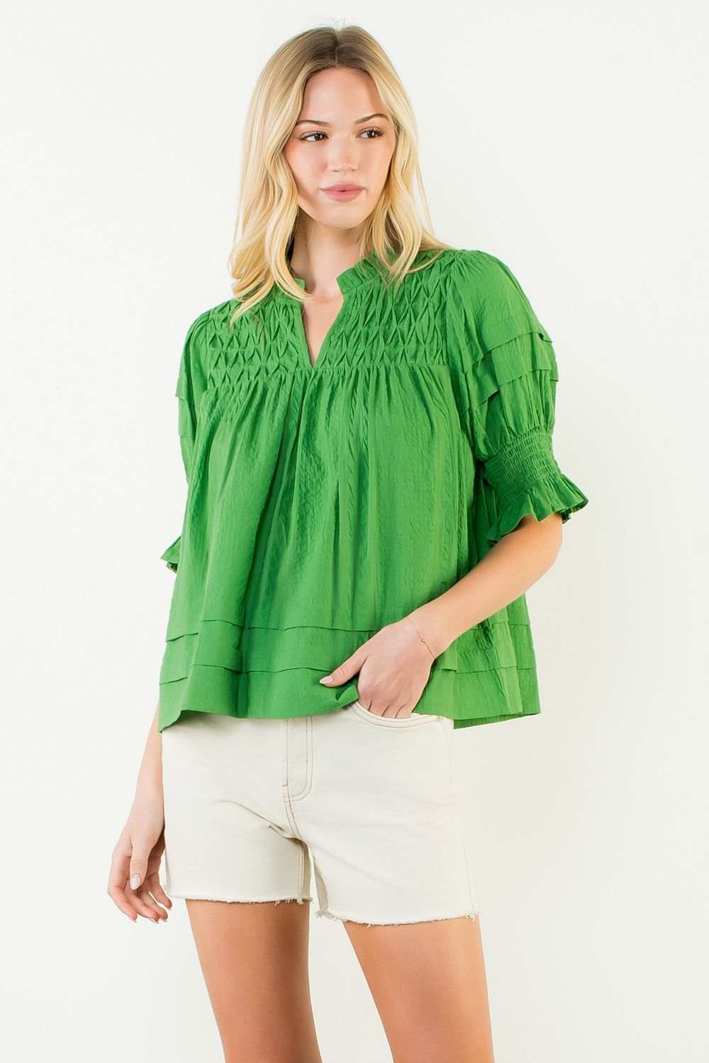 Woman wearing a green Alexandra short sleeve crinkle top with smocked detailing and white shorts.