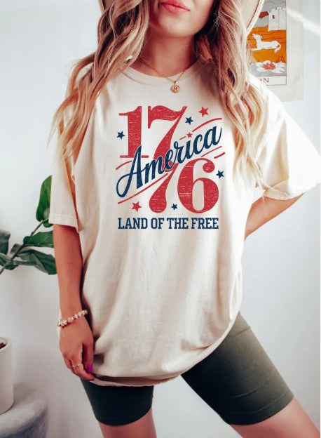 Woman wearing a natural-colored America 1776 Land of the Free t-shirt with olive green biker shorts.