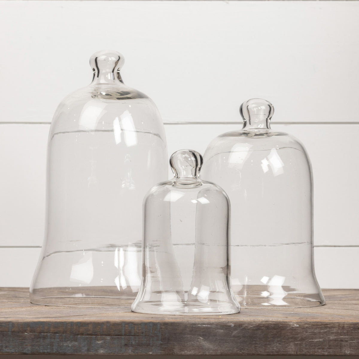 Three clear glass bell-shaped cloches in varying sizes on a rustic wooden surface.