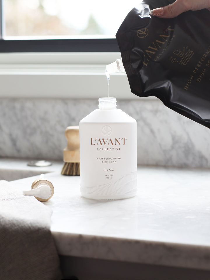 Pouring High Performing Dish Soap Refill into a white L'AVANT Collective bottle on a marble counter.