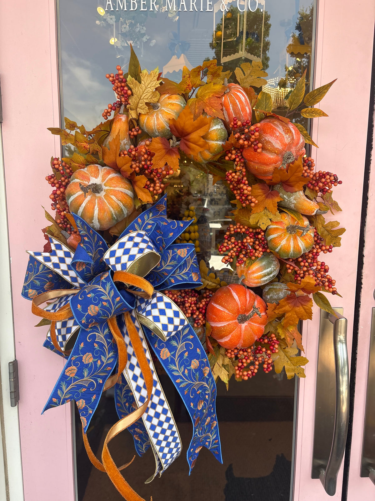 Handcrafted fall wreath with orange pumpkins, berries, and a large blue patterned bow on a pink door