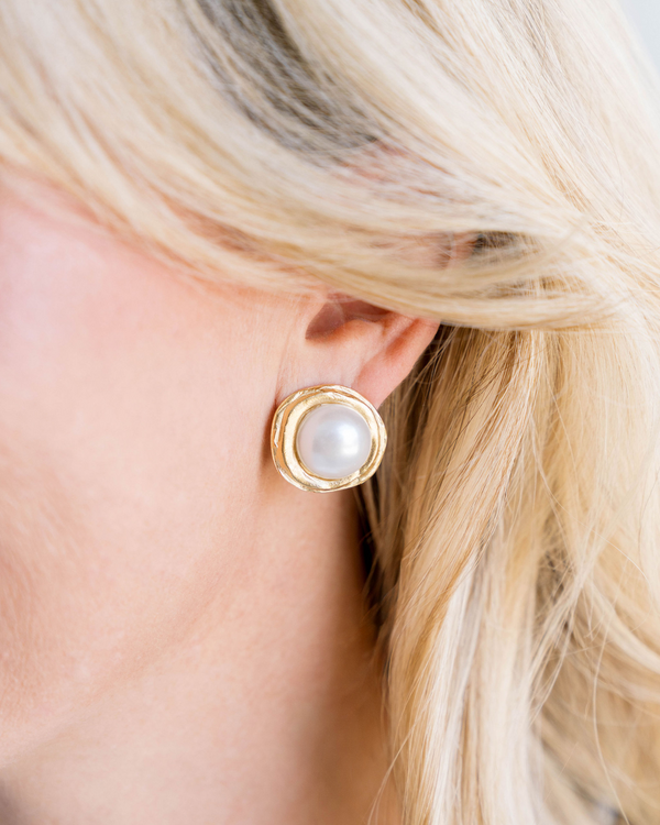 Close-up of a woman wearing a gold-rimmed pearl stud earring with a textured finish.