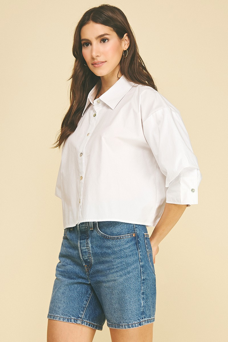 Woman wearing a white cropped button-down shirt with 3/4 tuck detailed sleeves and denim shorts.