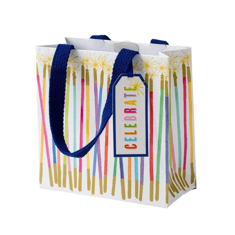Small square gift bag with a white background, decorated with colorful vertical stripes resembling candles and gold accents. A blue tag reads 'CELEBRATE'.