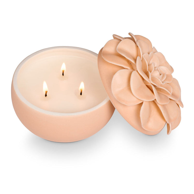 A peach-colored ceramic flower candle with three lit wicks and a decorative floral lid.