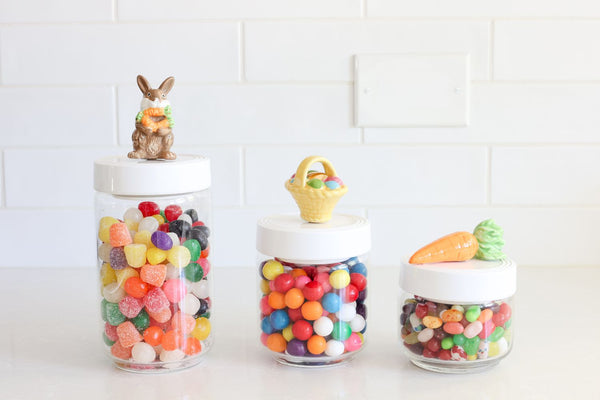 Three glass Pinstripes Canisters with white lids topped with a bunny, basket, and carrot figurine.