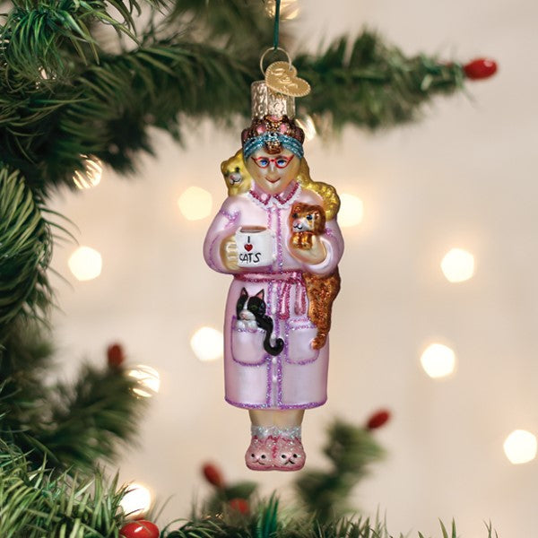 Glass cat lady ornament in a pink robe holding a mug, covered in cats, hanging on a Christmas tree.