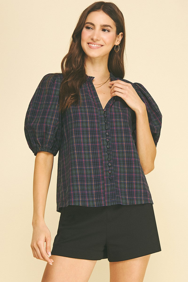 Woman wearing the April Plaid Puff Sleeve Blouse in a dark plaid pattern with black shorts.