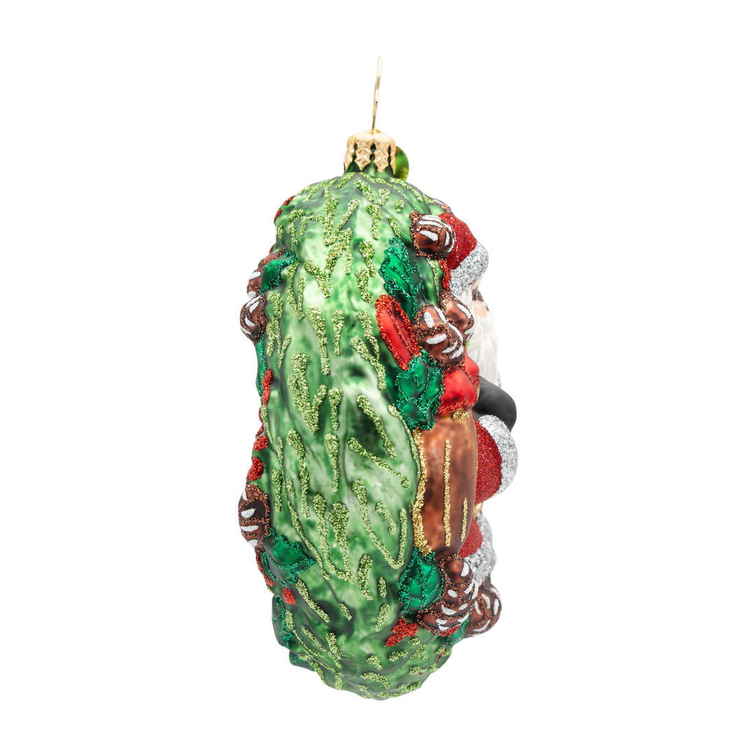 Side view of a Santa Claus glass ornament emerging from a green glittery wreath with pinecones.
