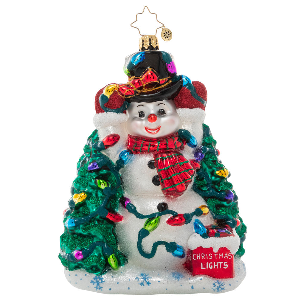 Glass Christmas ornament of a snowman wearing a hat and scarf, surrounded by trees and Christmas lights.