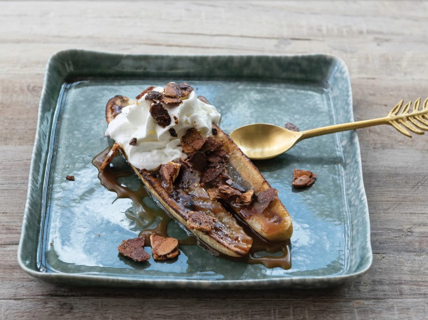 A split banana topped with whipped cream, chocolate shavings, and caramel sauce on a rectangular blue-green plate.