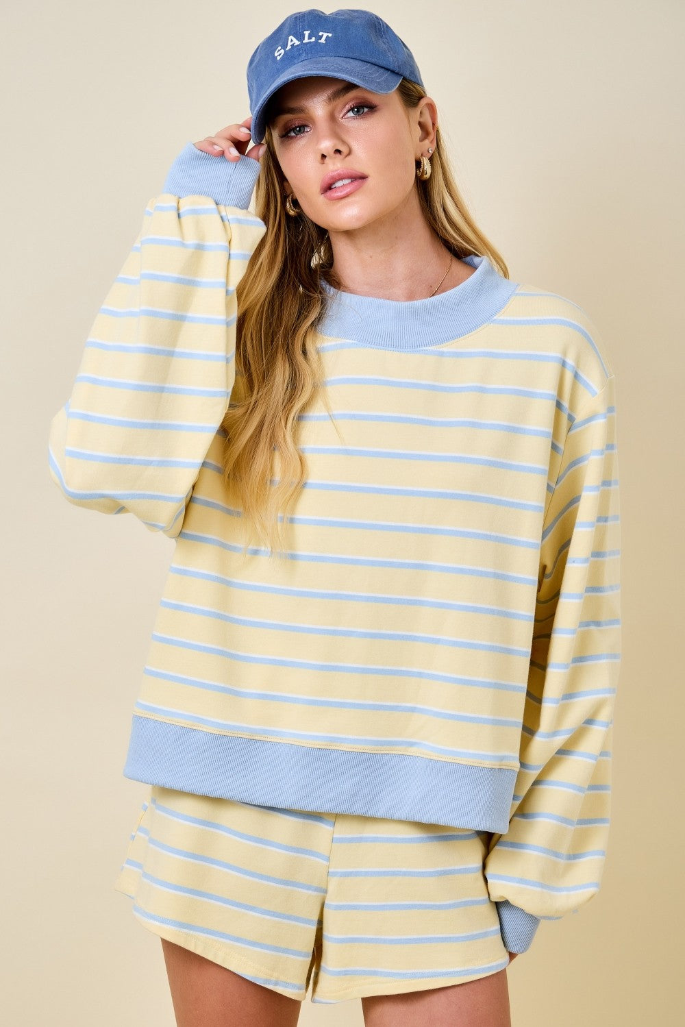 Woman wearing a yellow and blue striped mockneck sweatshirt with matching shorts and a blue cap.