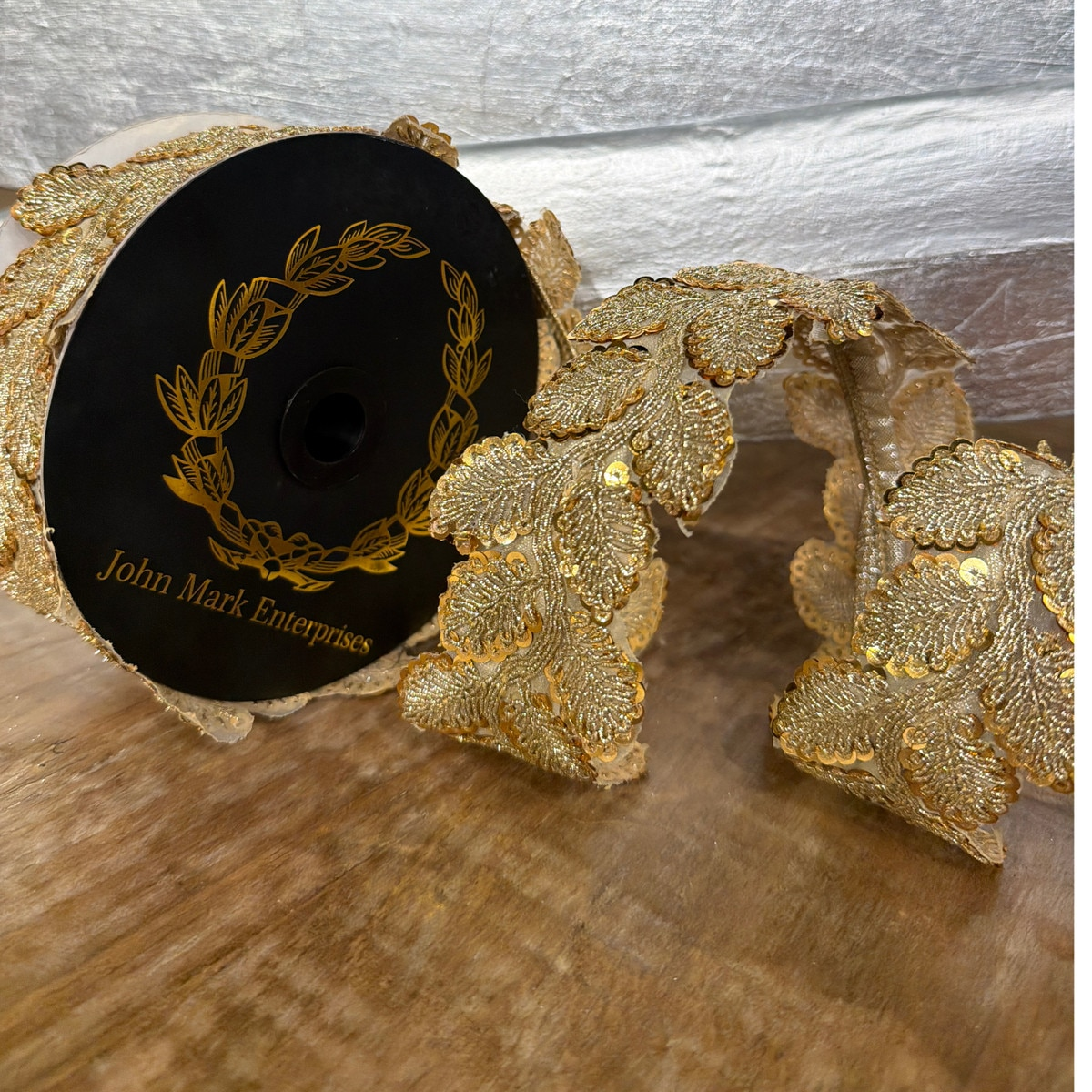 Spool of gold metallic sequin leaf trim ribbon by John Mark Enterprises on a wooden surface.