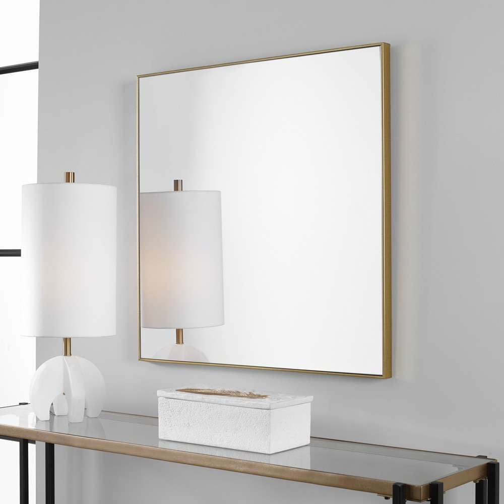 Alexo square wall mirror with a thin brushed gold frame mounted above a glass console table.