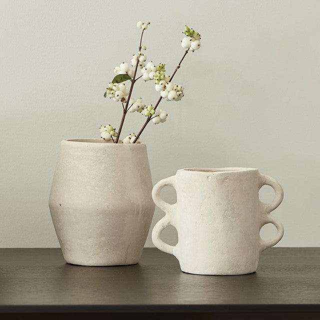 Two natural paper mache vases on a dark surface, one with four handles and one with white berries.