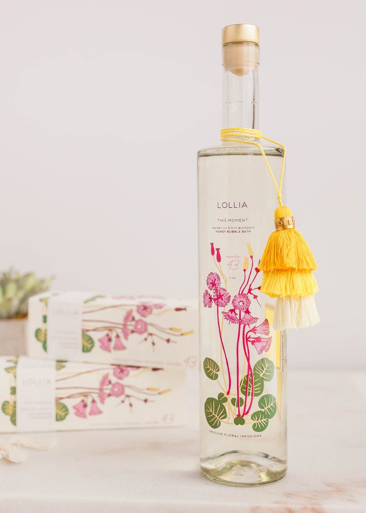 Slender glass bottle of This Moment Bubble Bath with tiered tassel and gold-accented cork cap.