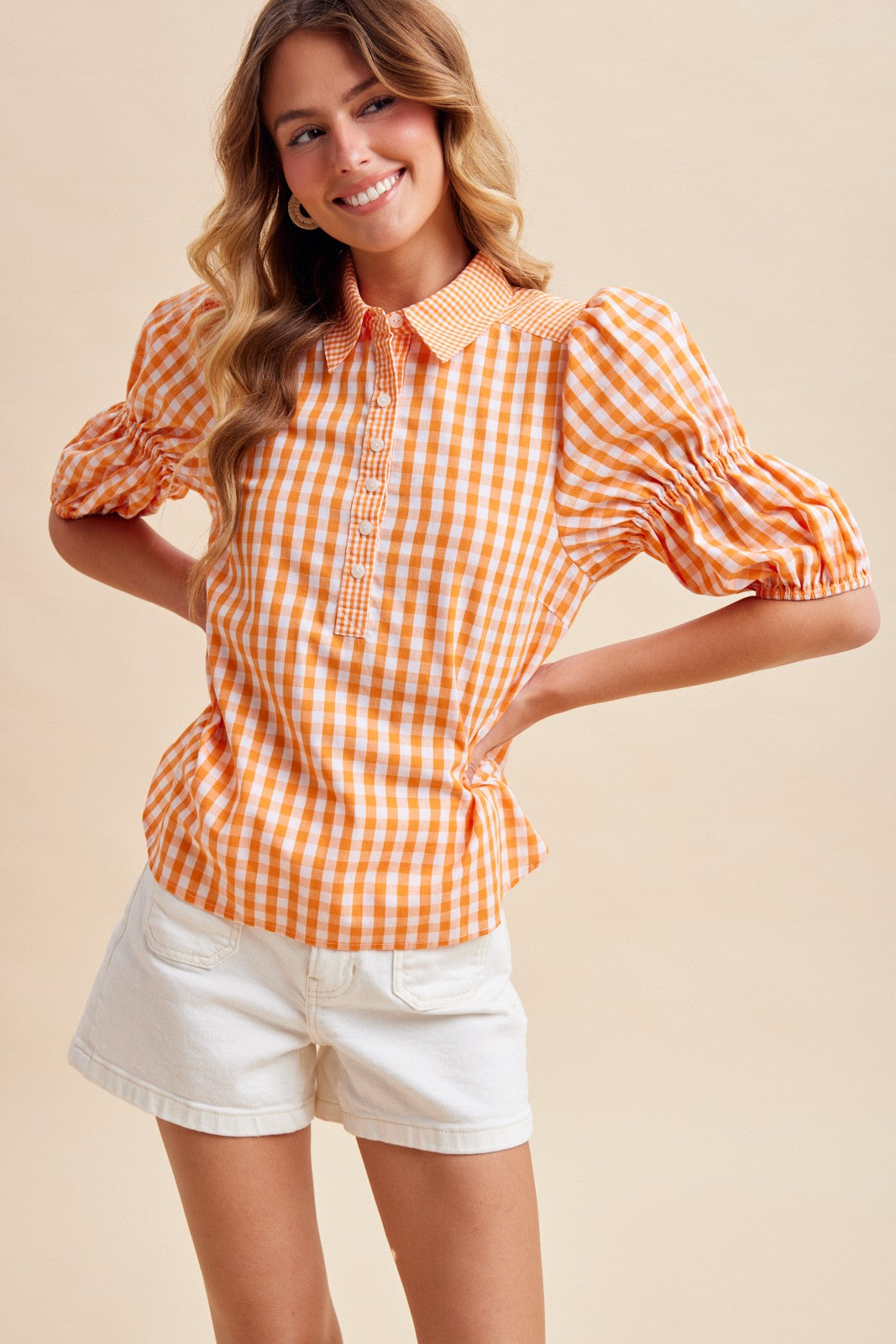 Woman wearing an orange and white Suzy Gameday Gingham Puff Sleeve Henley Blouse with white shorts.