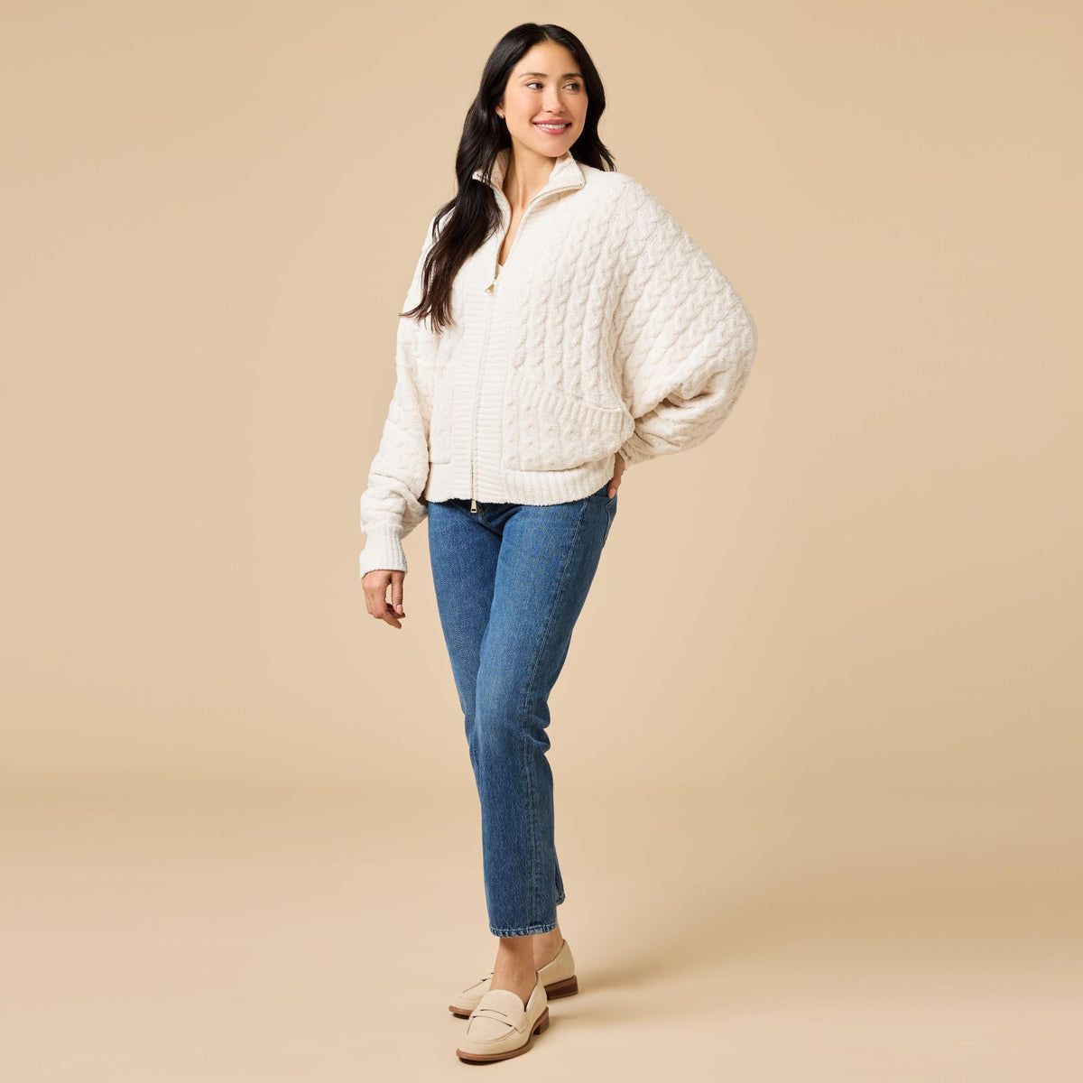 Woman in a cream cable knit funnel neck double zip jacket and blue jeans.