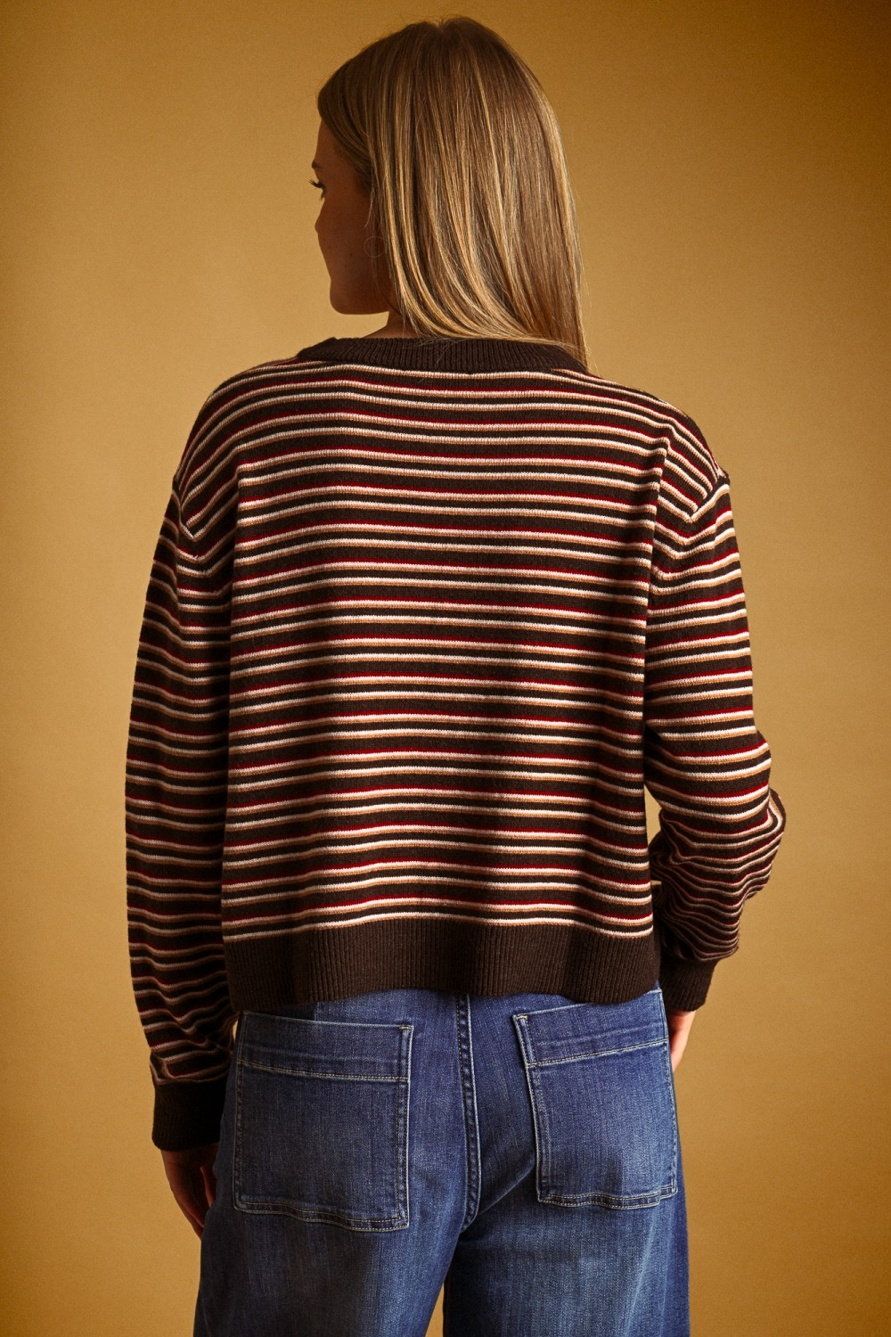 Back view of a woman wearing a multi-color striped knit top with brown ribbed trim and blue jeans.
