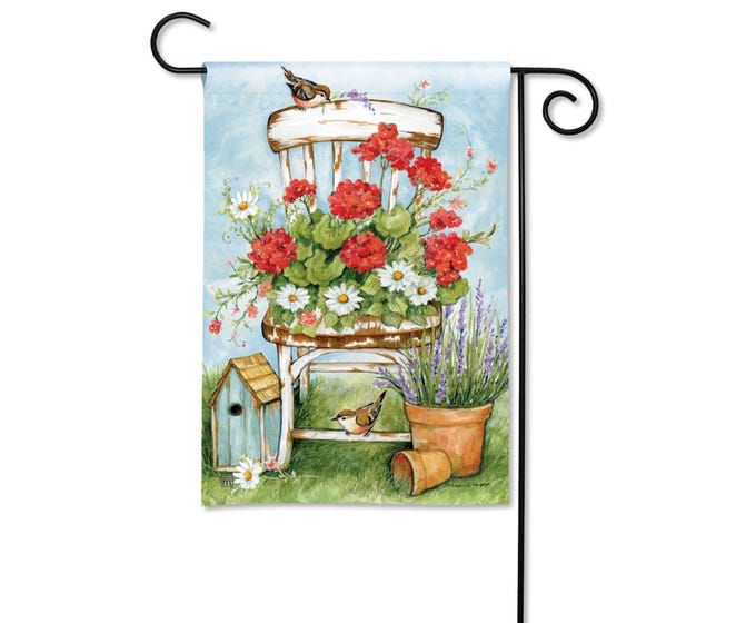 Garden flag with a watercolor painting of a white chair overflowing with red geraniums and daisies, a birdhouse, and lavender.