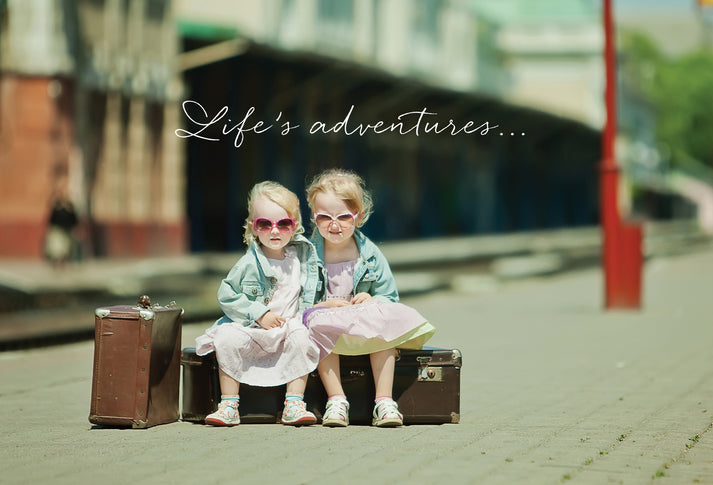 Greeting card with cursive text 'Life's adventures...' above two young girls in sunglasses and jackets sitting on suitcases.