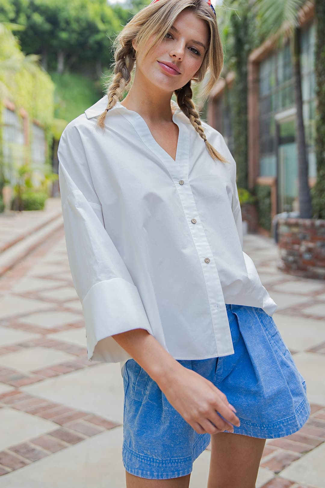 Woman wearing a white Blake Back Pleat Button Down Blouse with wide sleeves and blue shorts.