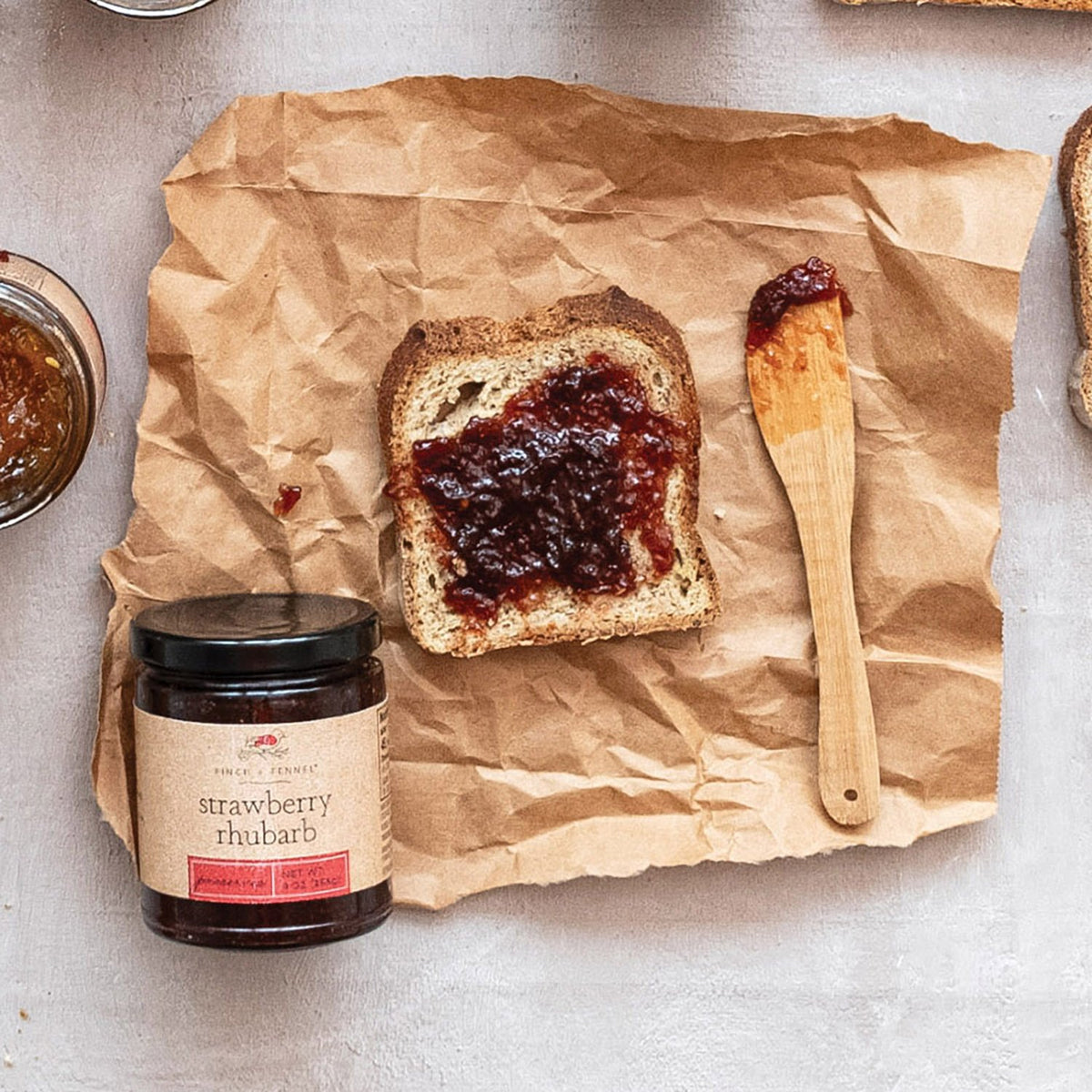 A jar of Strawberry Rhubarb Preserves next to a slice of toast spread with jam on brown paper.
