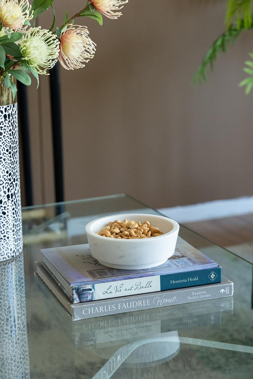White marble bowl filled with nuts, resting on a stack of books on a glass coffee table.