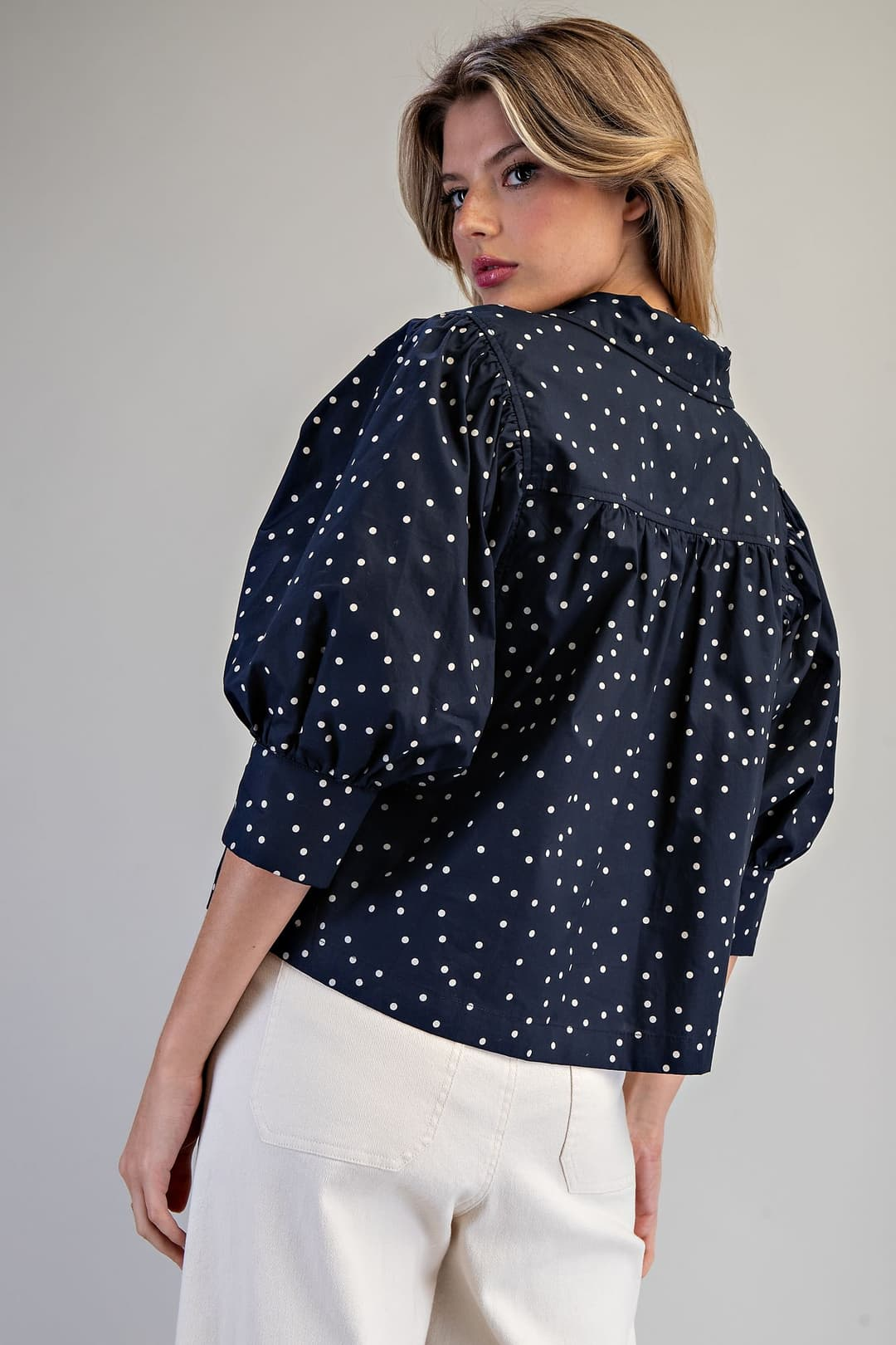 Back view of a woman wearing the Molly navy blue polka dot blouse with puff sleeves and white pants.