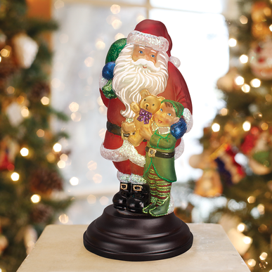 Santa figurine with an elf holding a teddy bear, on a dark base, with a festive background.