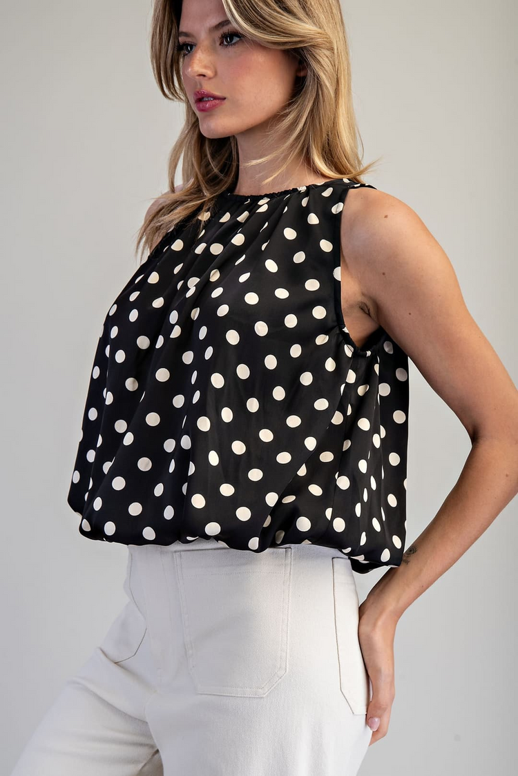 Woman wearing the Monique Polka Dot Sleeveless Blouse in black with white dots and white trousers.