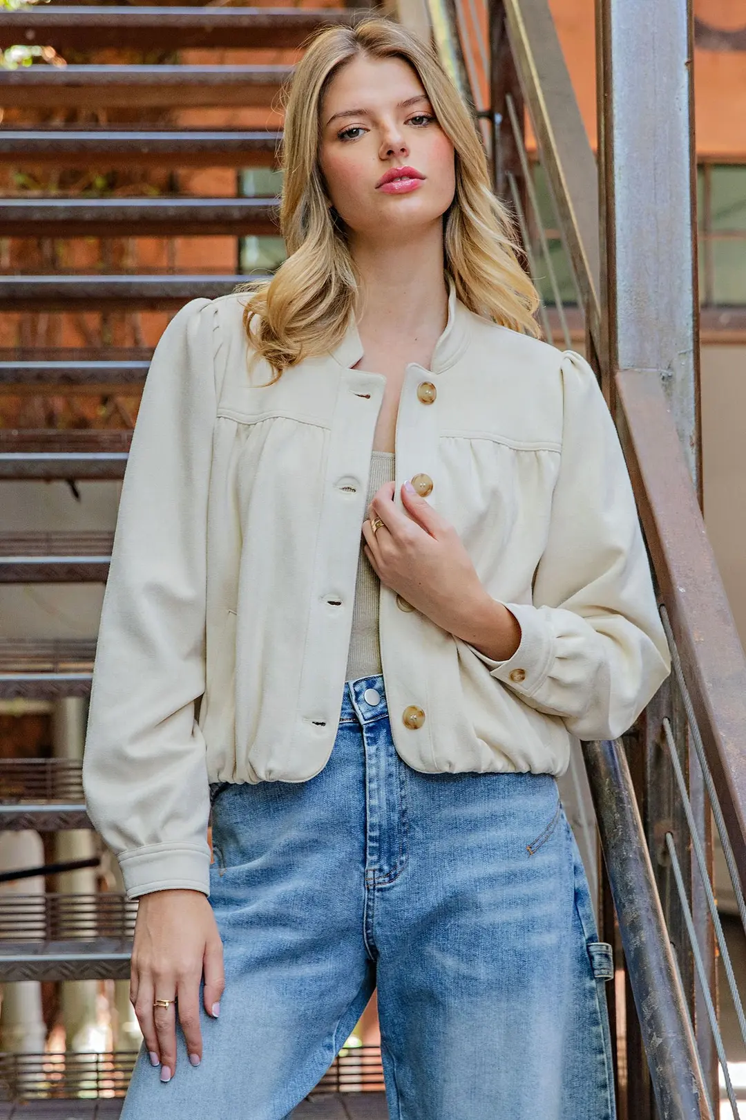 Woman wearing a cream Bette Suede Blouson Hem Button Up Jacket with blue jeans on a staircase.