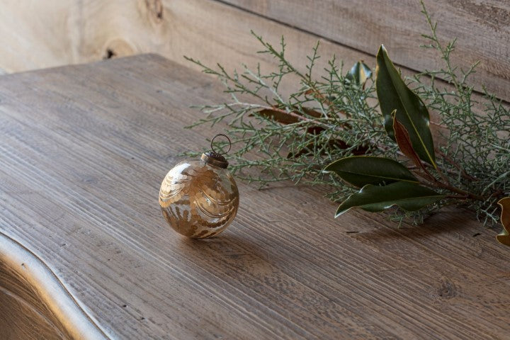 Etched Golden Garland Glass 3" Ornament on a rustic wooden surface next to evergreen branches.