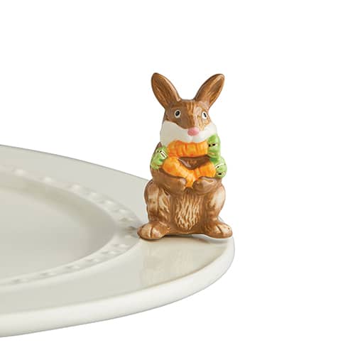 Brown ceramic bunny figurine holding carrots and green sprouts on a white plate.