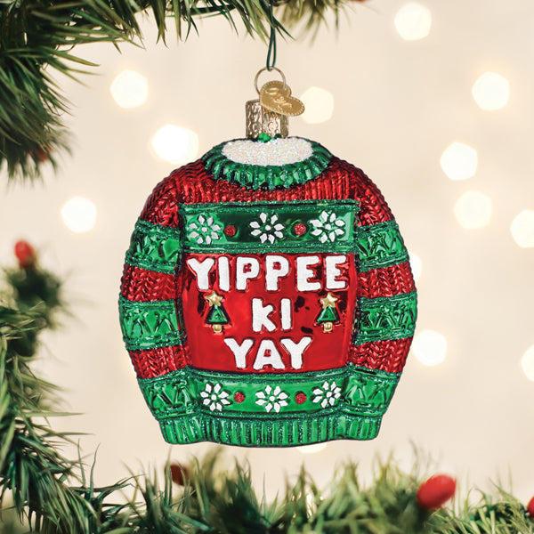 Red and green glass sweater ornament with white 'Yippee Ki Yay' text hanging on a Christmas tree.