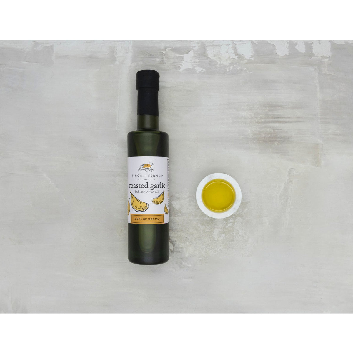 A 6.8 oz bottle of Finch + Fennel Roasted Garlic Infused Olive Oil next to a small bowl of oil.