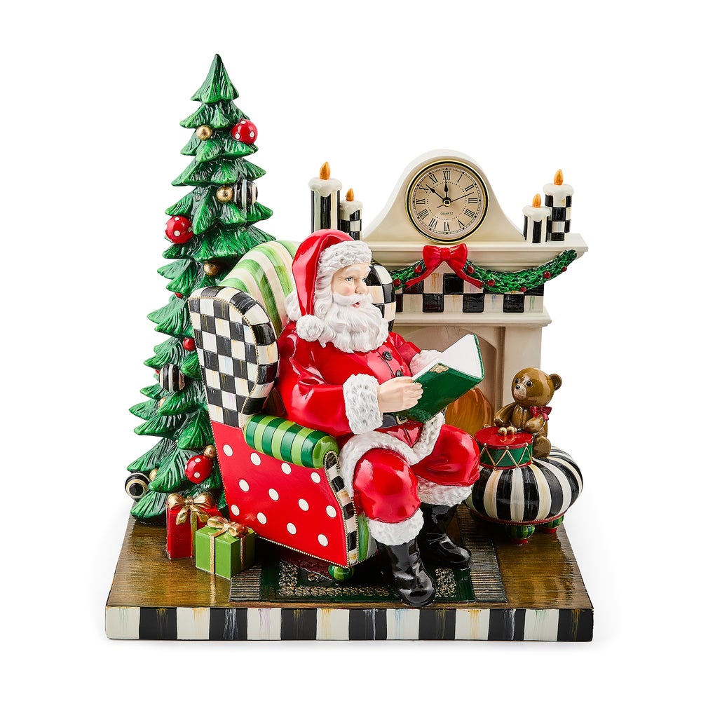 Santa figurine sitting in a patterned armchair reading a book by a fireplace and Christmas tree.