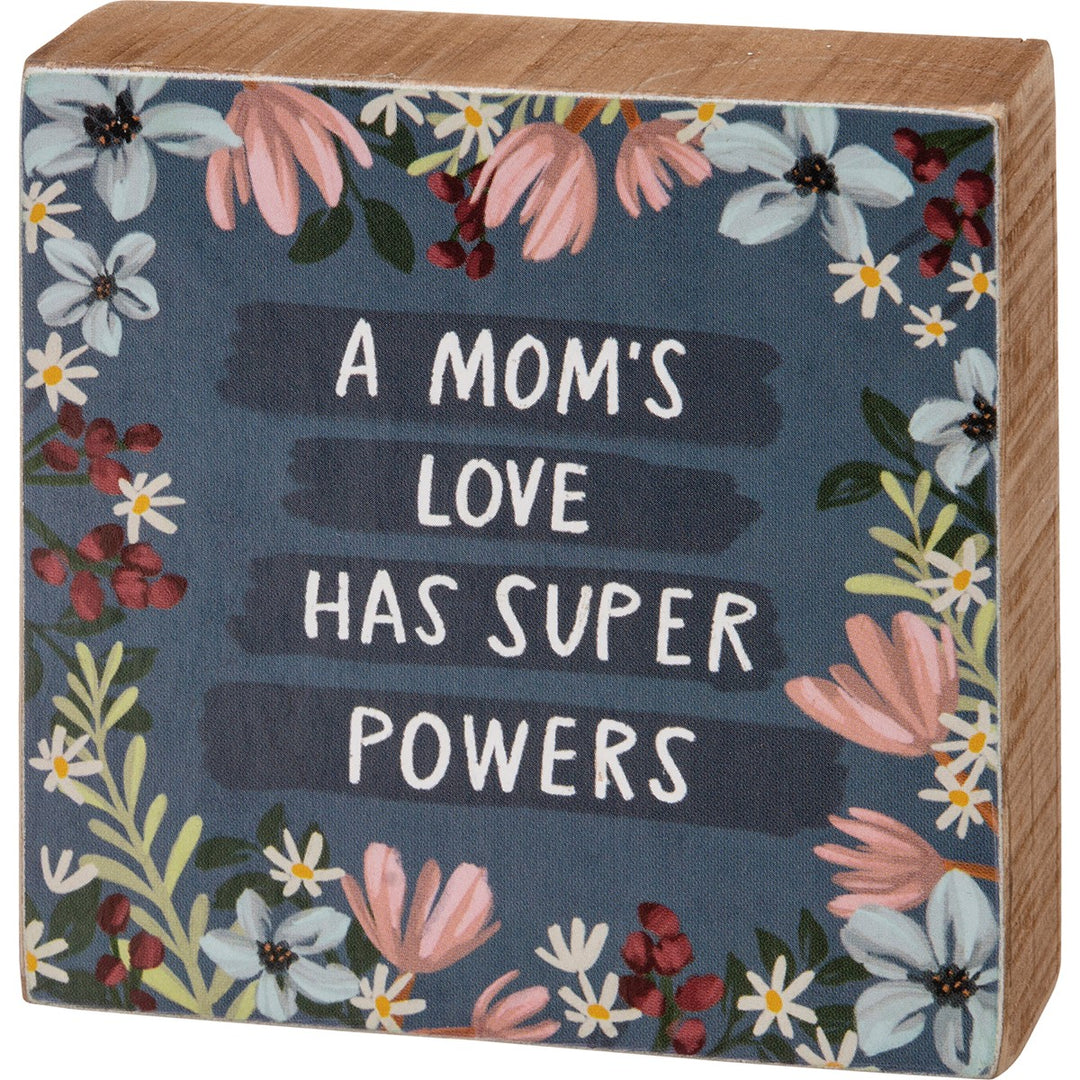 Wooden block sign with floral border and text 'A MOM'S LOVE HAS SUPER POWERS' on a dark blue background.