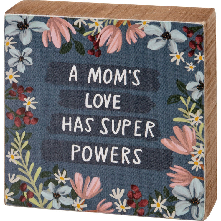 Wooden block sign with floral border and text 'A MOM'S LOVE HAS SUPER POWERS' on a dark blue background.