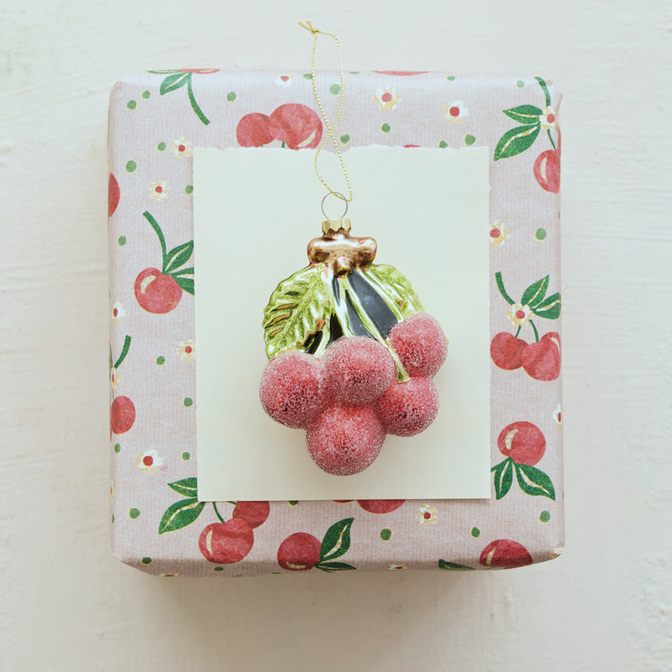 Red sugared glass cherry cluster ornament with green leaves on a cherry-patterned gift box.