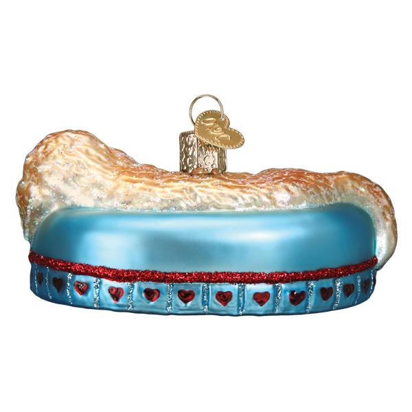 Glass Sleepy Doodle Dog ornament lying on a blue bed with red glitter hearts and a gold heart tag.