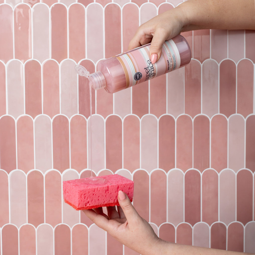 Sweet Grace Dish Soap being poured onto a pink sponge against a pink tiled wall.